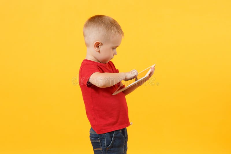 Little cute kid baby boy 3-4 years old in red t-shirt holding in hand tablet pc computer isolated on yellow background. Kids childhood lifestyle concept. Problem of children and gadgets. Copy space. Little cute kid baby boy 3-4 years old in red t-shirt holding in hand tablet pc computer isolated on yellow background. Kids childhood lifestyle concept. Problem of children and gadgets. Copy space